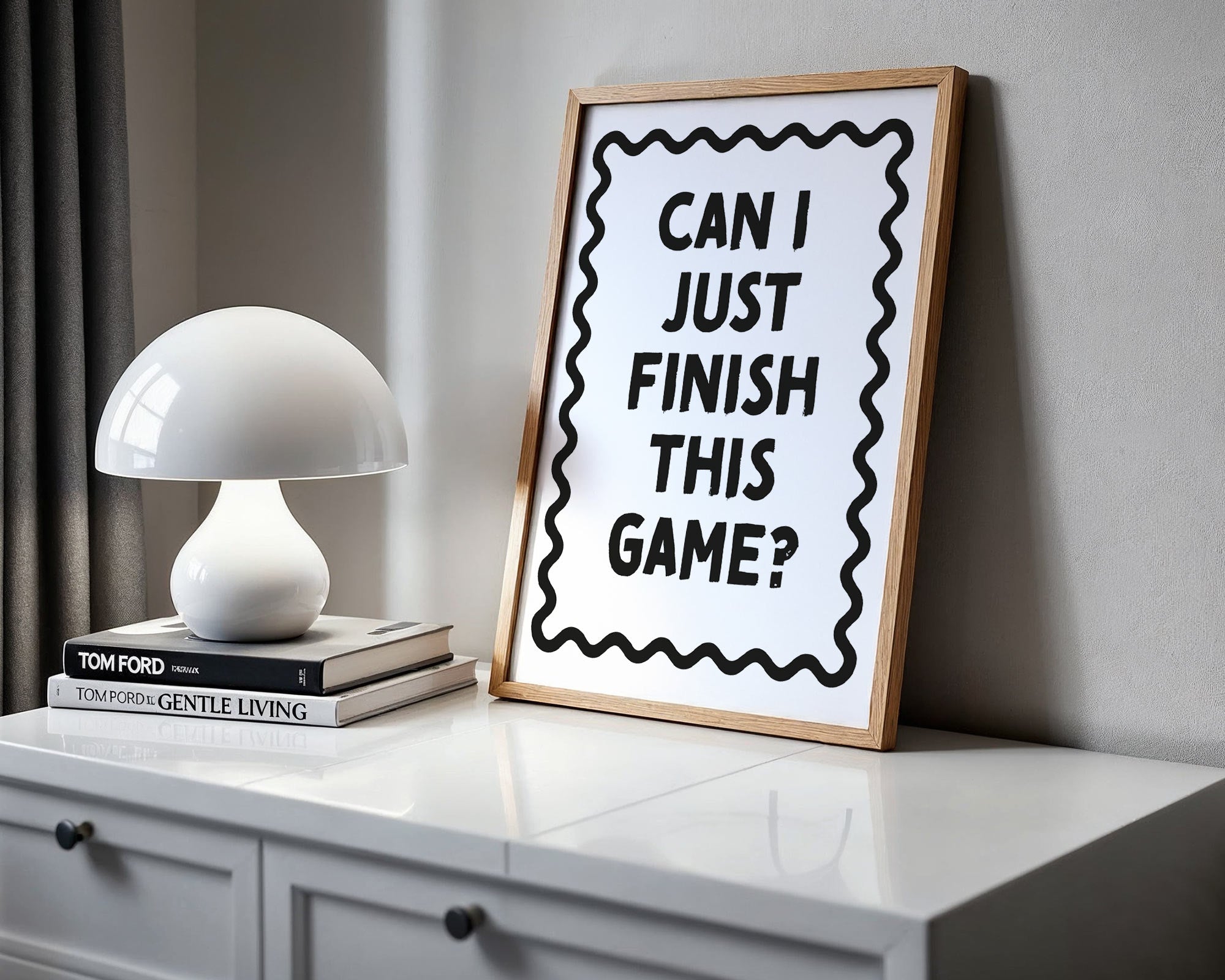 Framed poster with text 'Can I Just Finish This Game?' on a shelf with books and a lamp.