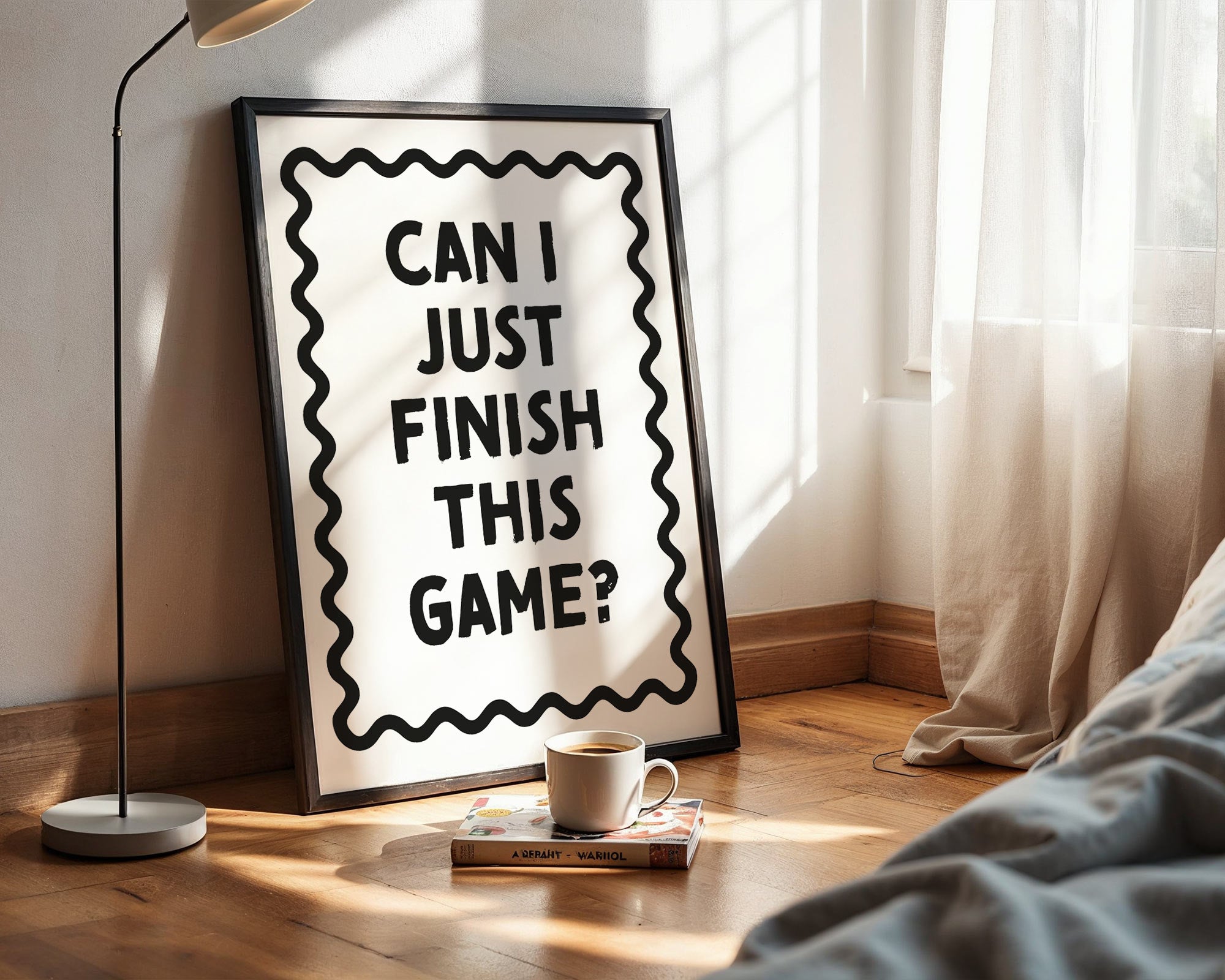Framed poster with text 'Can I just finish this game?' on a wooden floor next to a lamp and a cup.