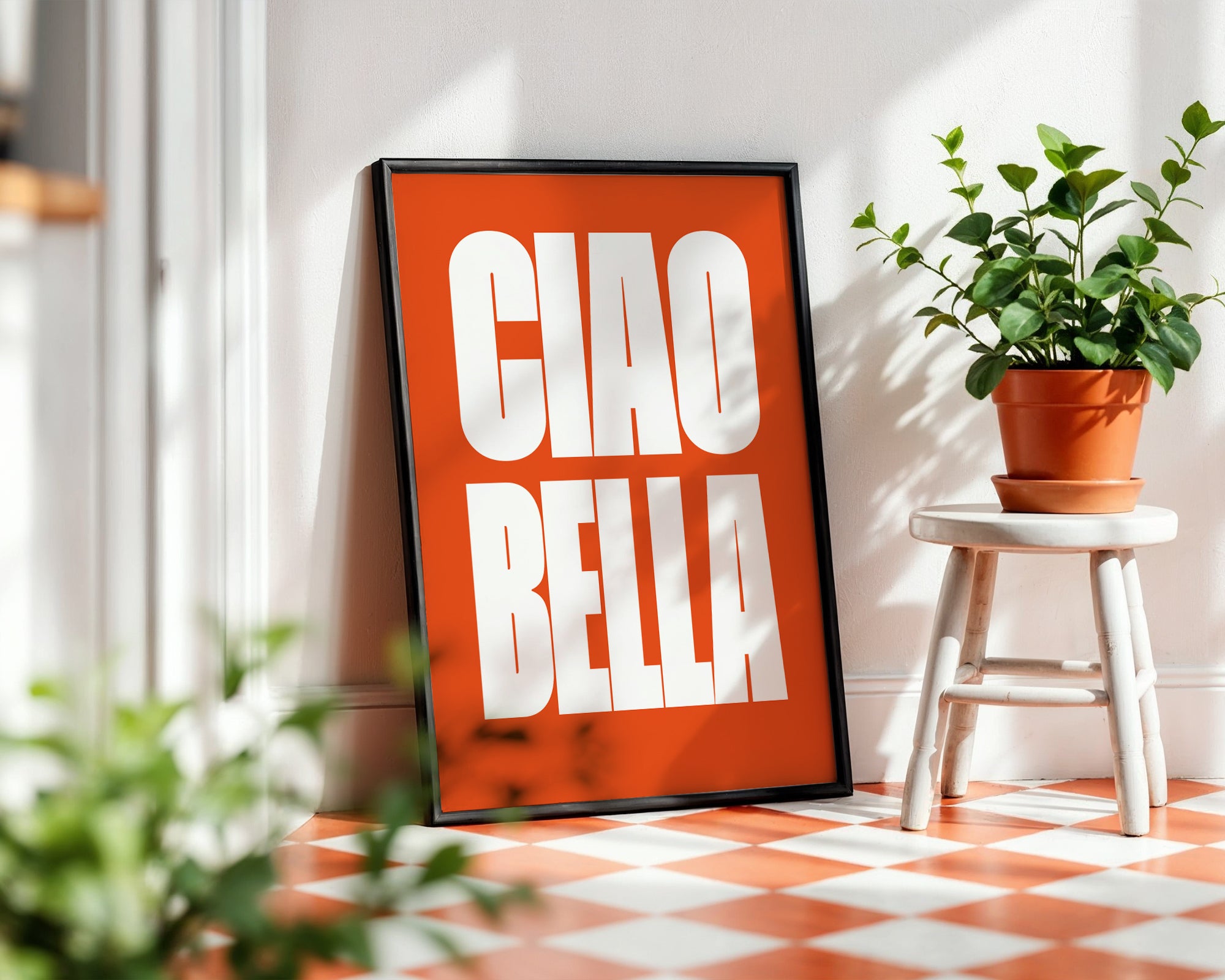 Framed artwork with 'CIAO BELLA' on an orange background in a room with plants and a checkered floor.