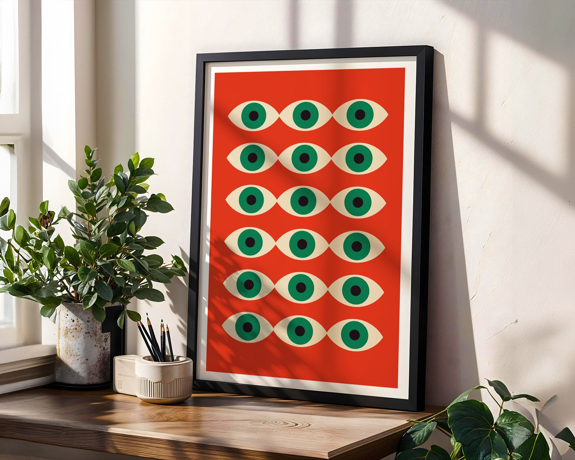 Framed artwork with eye pattern on a red background, placed on a wooden surface with plants.