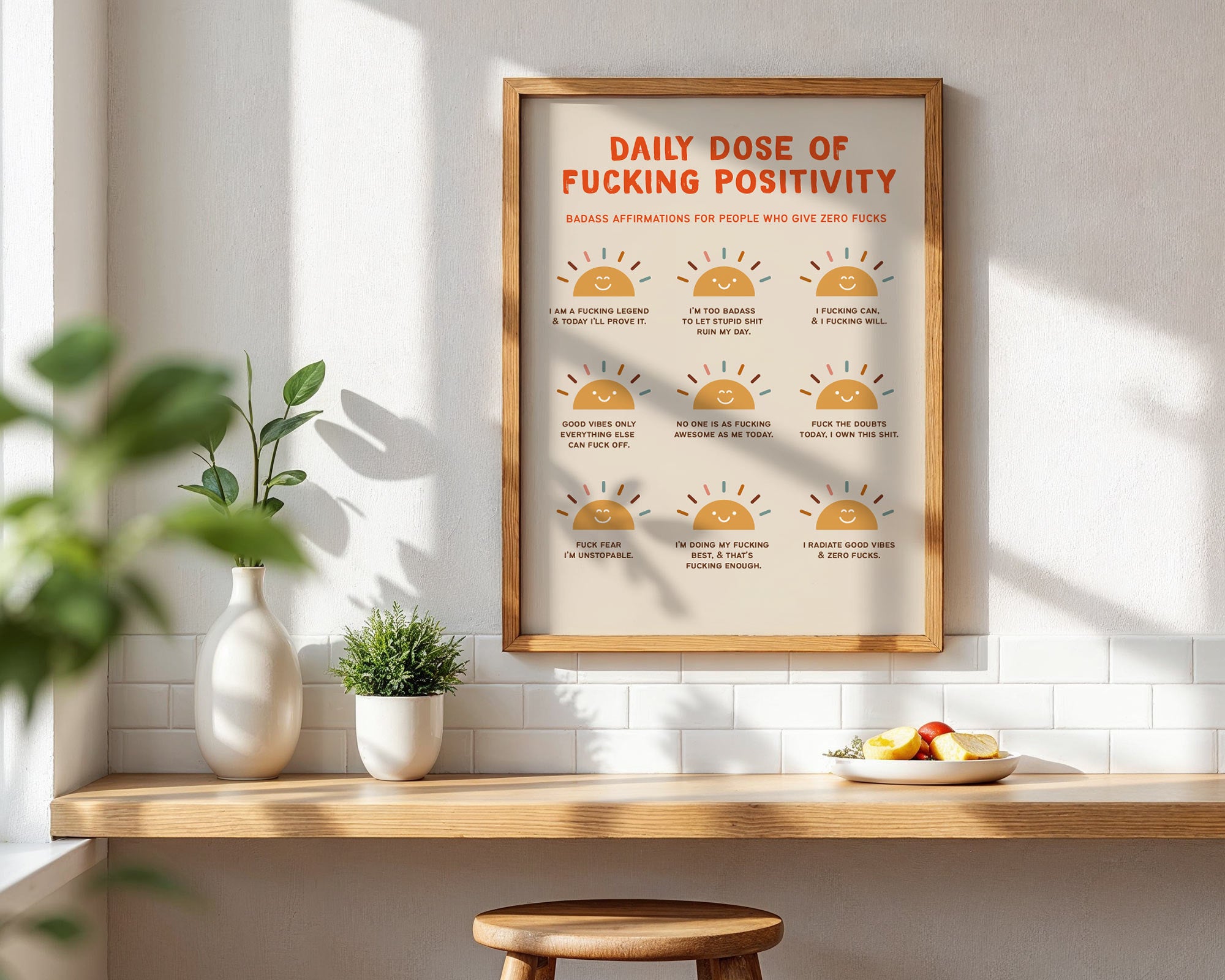Framed poster titled 'Daily Dose of Fucking Positivity' on a wall with a table and plants below.