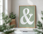 Framed poster with a large white ampersand and 'Chill' text on a green background, placed on a windowsill with plants.