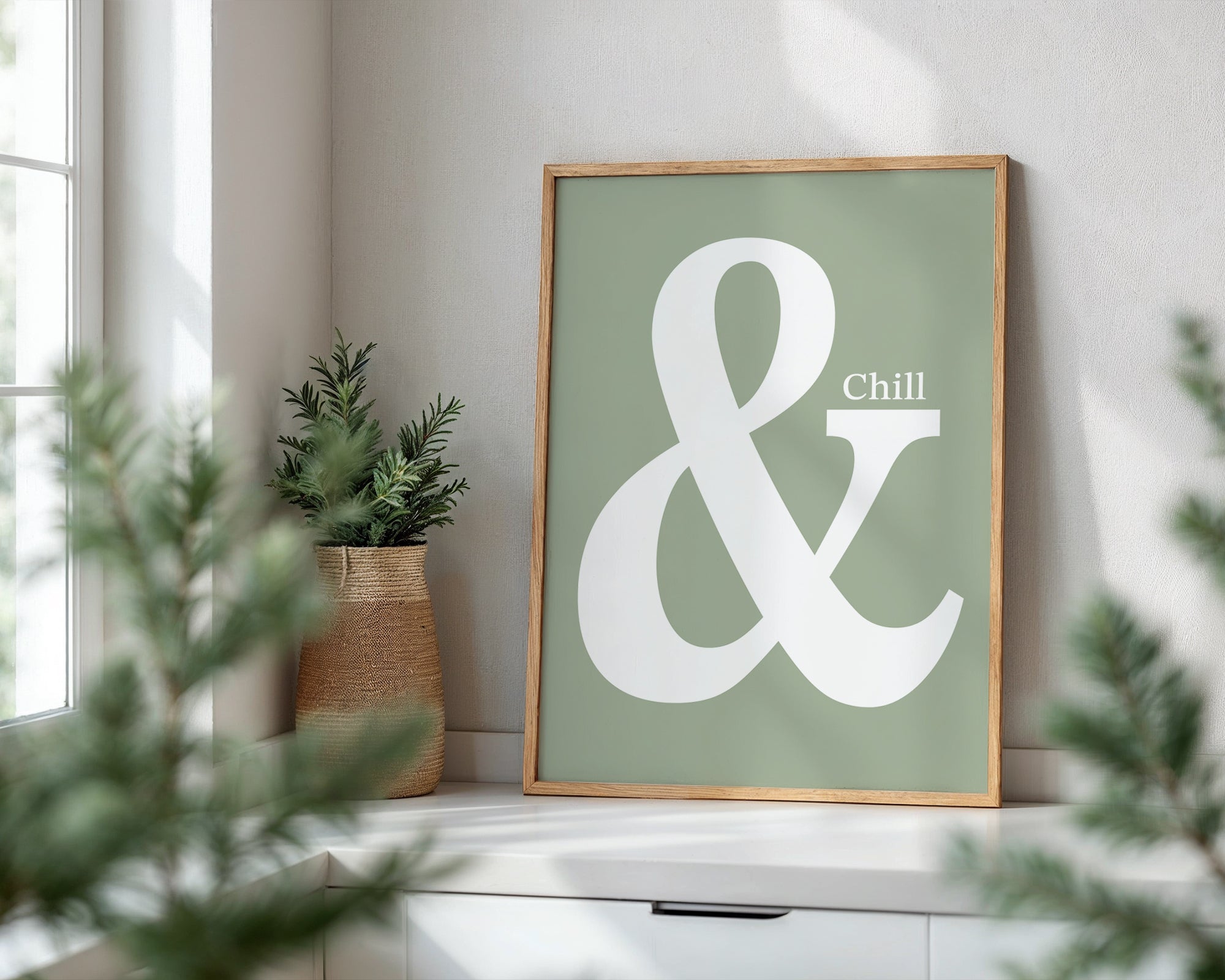 Framed poster with a large white ampersand and 'Chill' text on a green background, placed on a windowsill with plants.