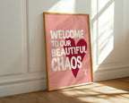 Framed poster with 'Welcome to our beautiful chaos' text on a pink background, leaning against a white wall.