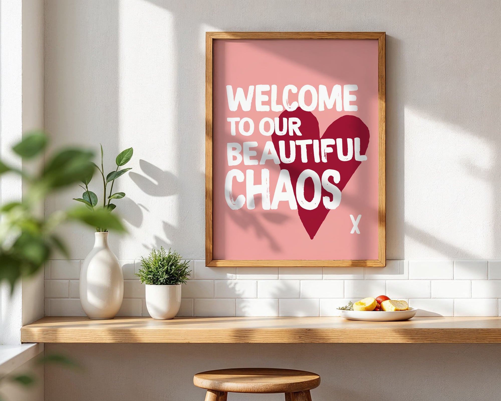 Framed artwork with 'Welcome to our beautiful chaos' text on a wall with a table and plants below.