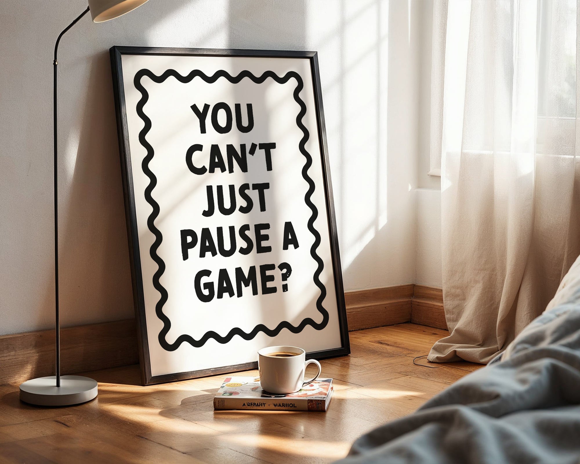 Framed poster with 'You can't just pause a game?' text in a room with a lamp and book.