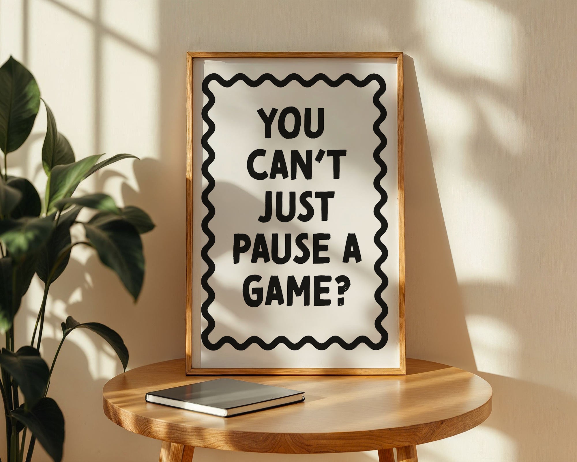 Framed poster with text 'You can't just pause a game?' on a wooden table with a plant in the background.