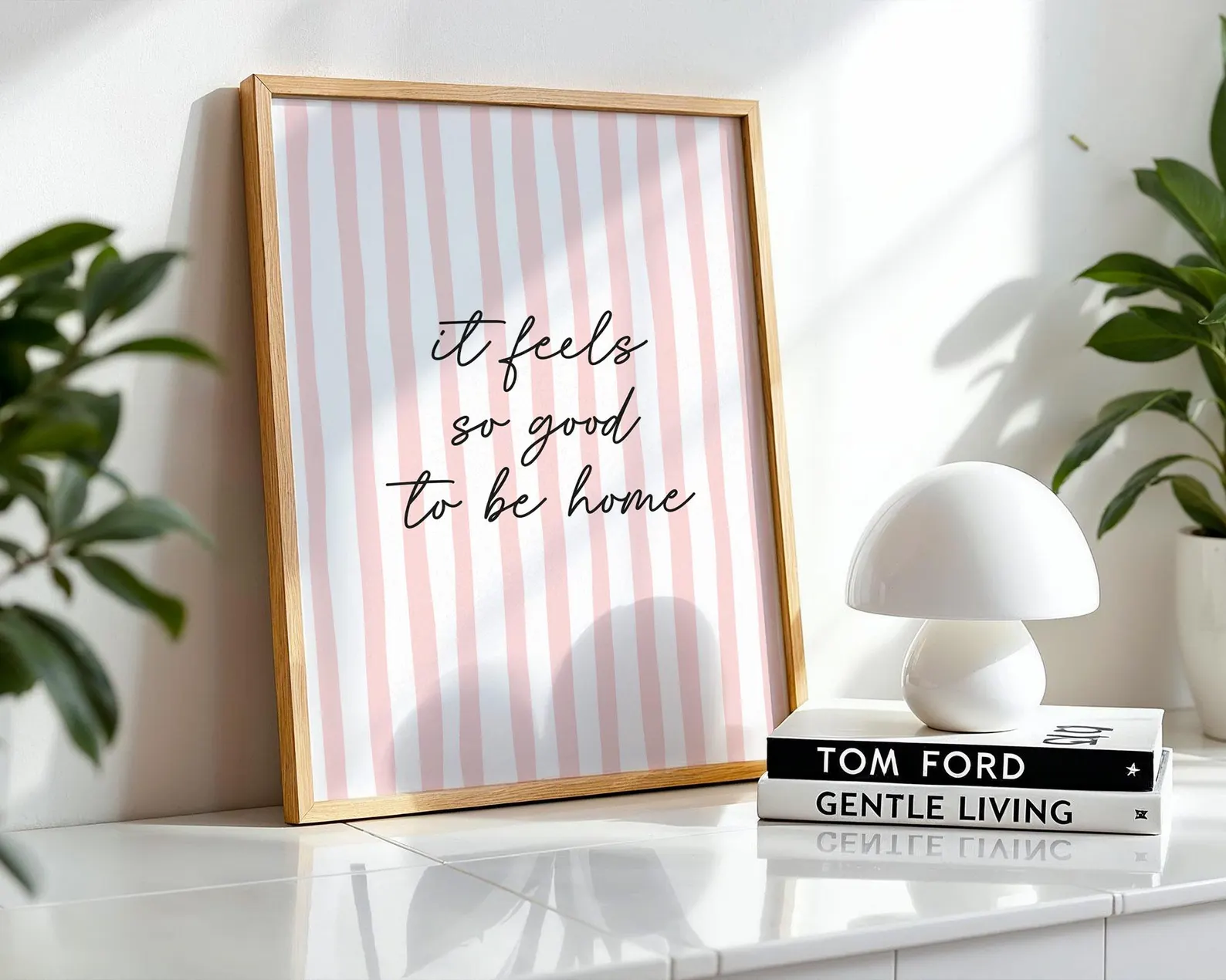 Framed print with 'it feels so good to be home' text on a striped background, placed on a surface with books and a lamp.