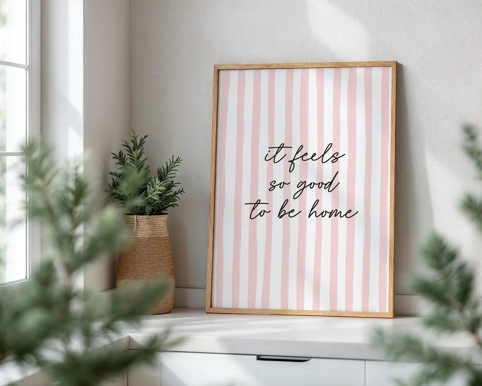 Framed print with 'it feels so good to be home' text on a striped background, placed on a windowsill with plants.