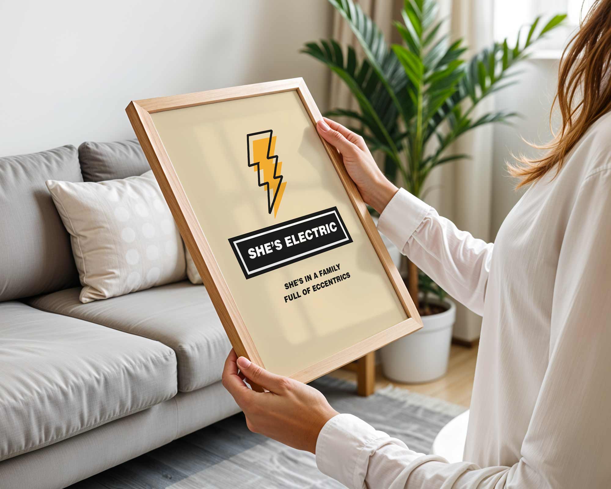 Person holding a framed sign with 'SHE'S ELECTRIC' text and logo in a living room.