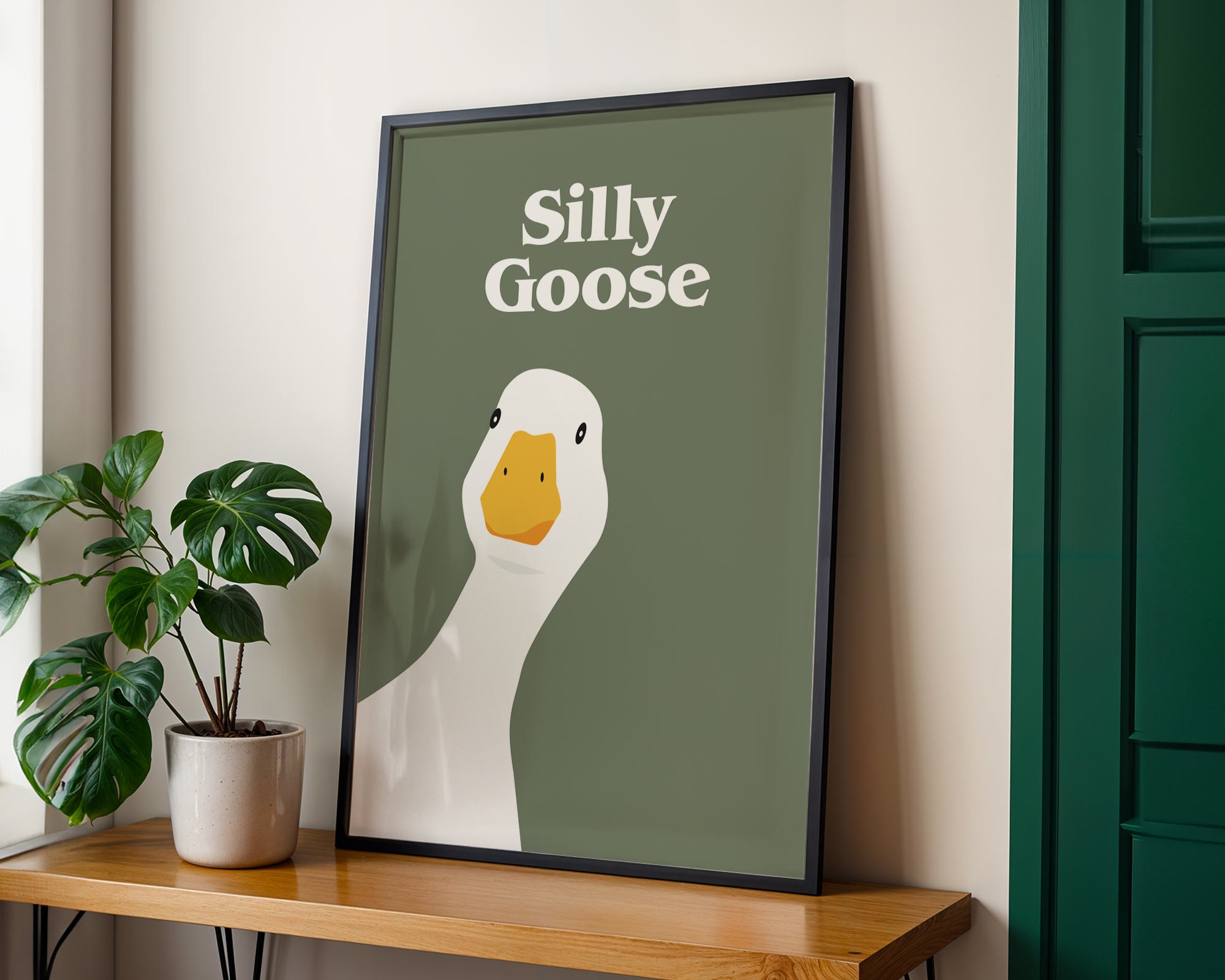 Framed artwork with a goose illustration and text 'Silly Goose' on a wall next to a plant.