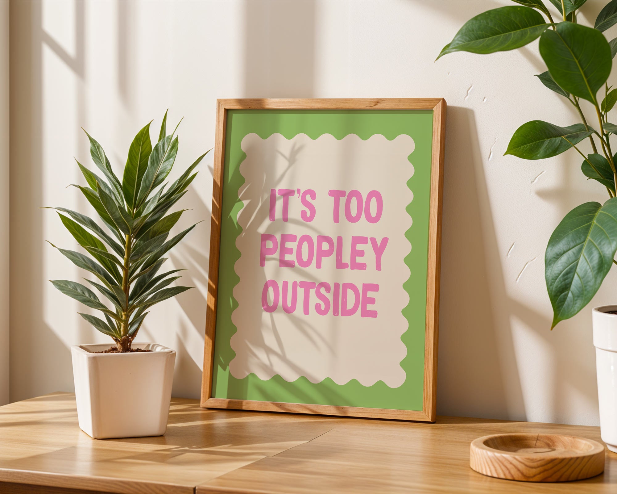 Framed sign with 'It's too peopley outside' text on a wooden surface with plants.