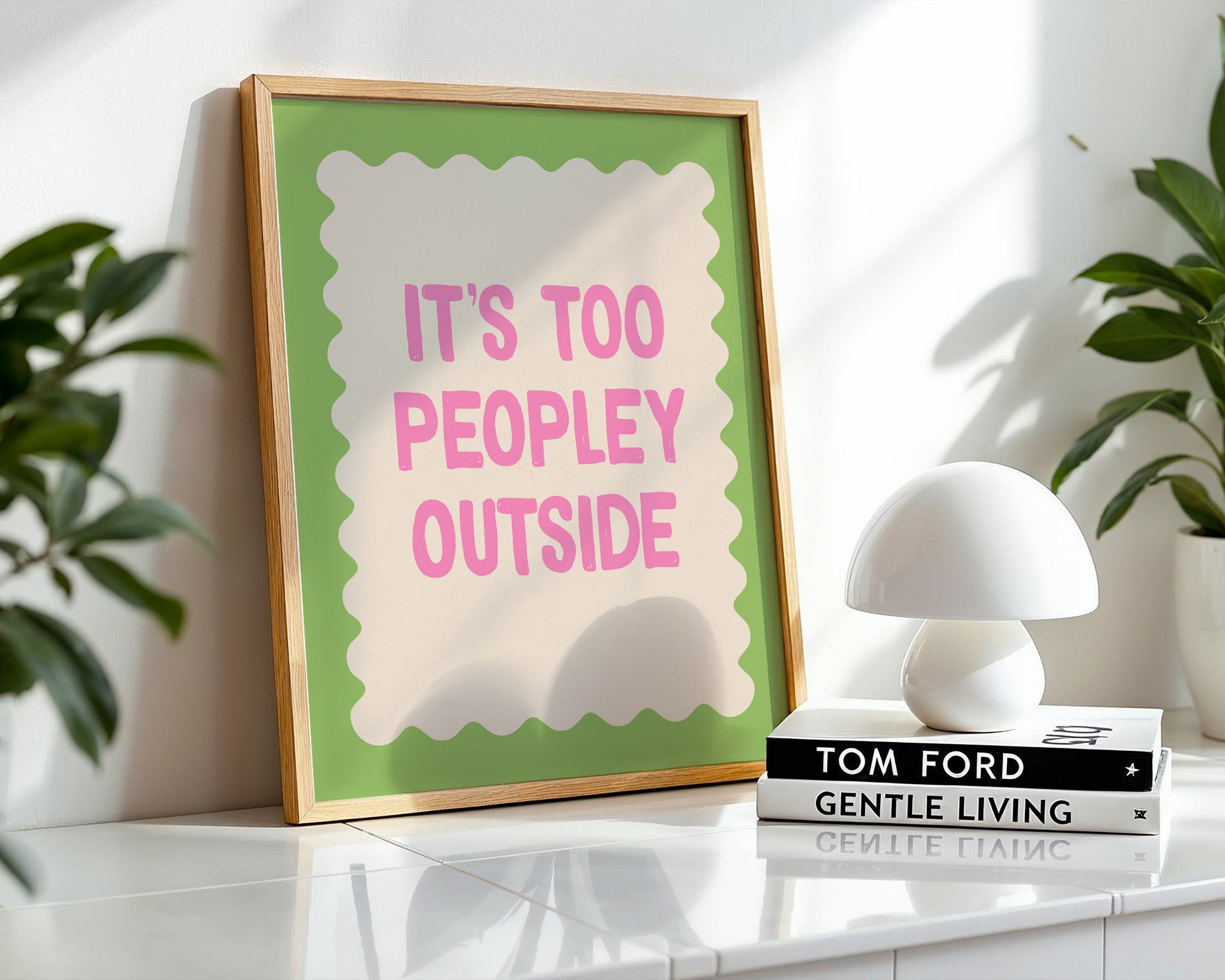 Framed print with 'It's too peopley outside' text on a shelf with books and a lamp.