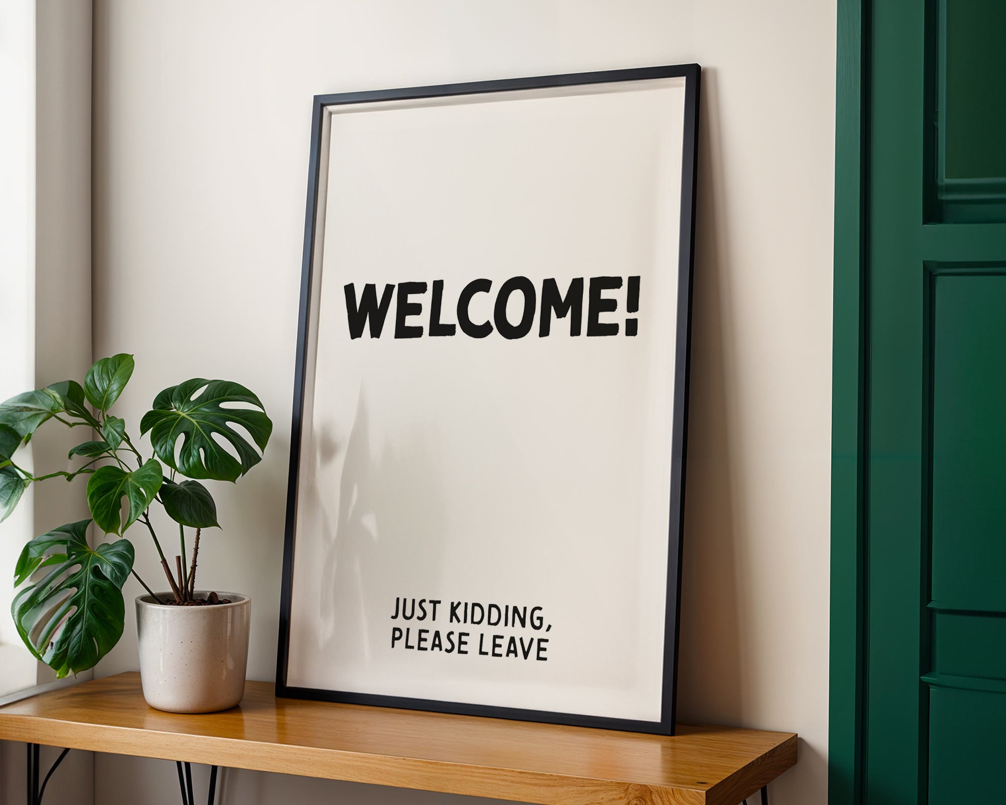 Framed poster with 'WELCOME! JUST KIDDING, PLEASE LEAVE' text on a wall next to a plant and wooden shelf.