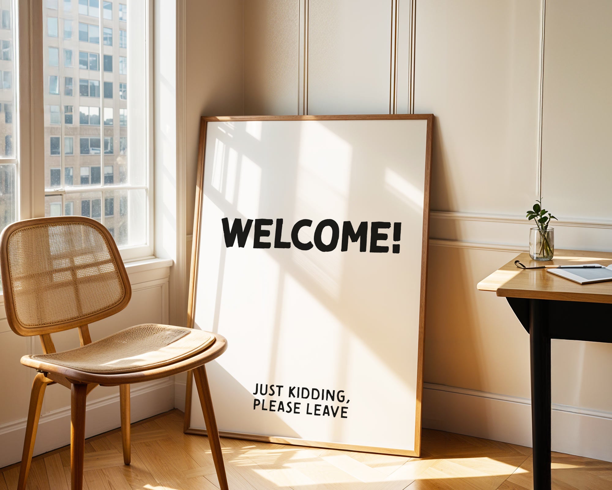 Framed sign with 'WELCOME! JUST KIDDING, PLEASE LEAVE' text in a room with a chair and window.